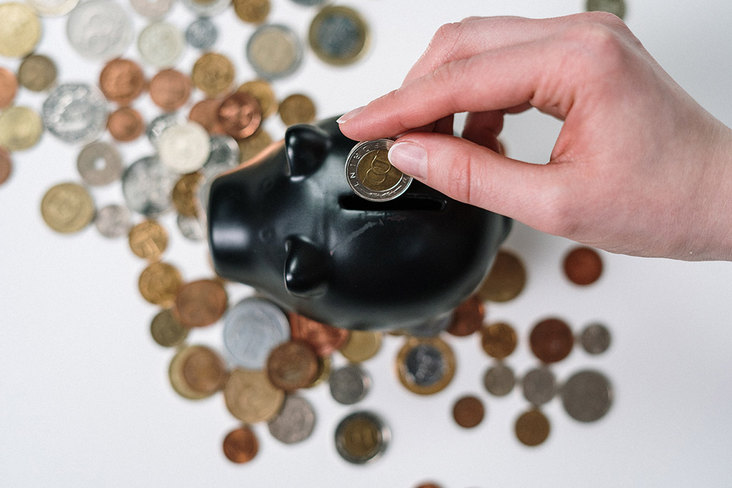 Top view of a black piggy bank with a lot of coins scattered around it and a hand