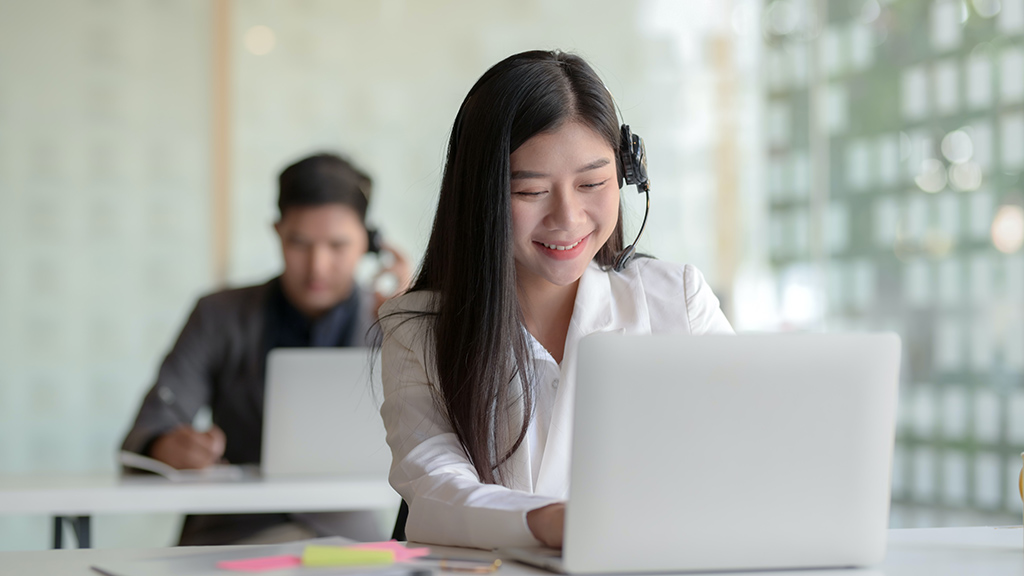 A smiling woman wearing a headset sitting on a table with an open laptop and passionately talking to a customer